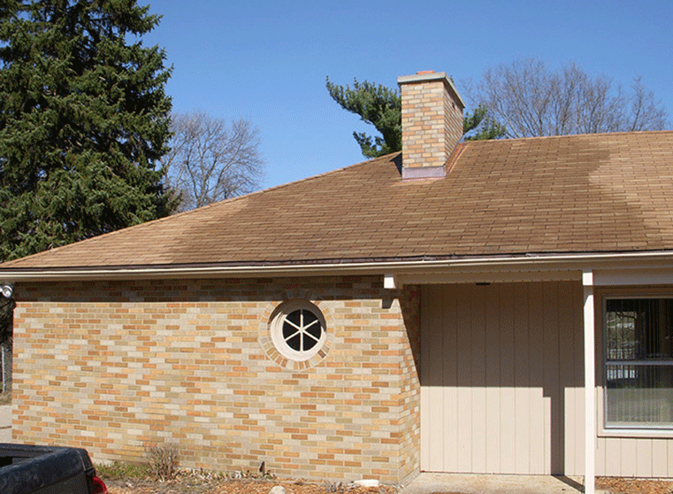 A tan brick house exterior features a round window, a brick chimney, and a brown shingled roof under a clear blue sky.