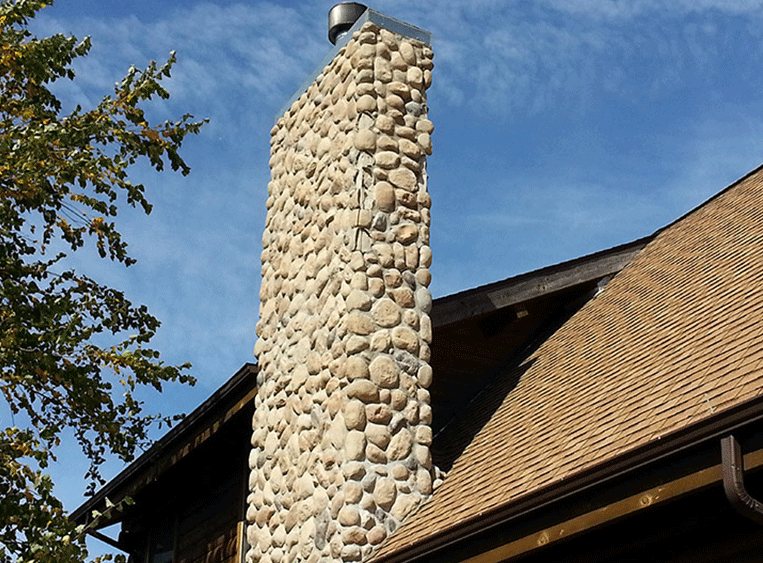 A tall stone chimney made of rounded, beige river rocks extends above the shingled roof of a house against a blue sky.