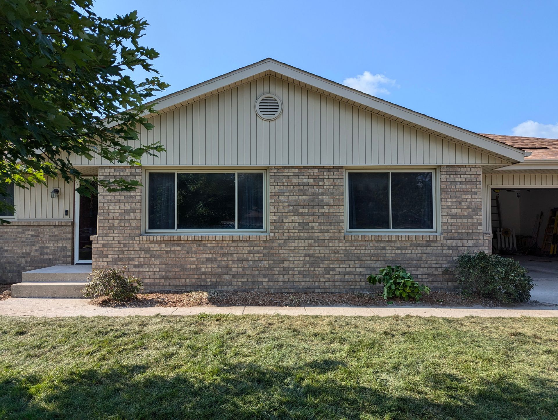 A single-story tan brick house with vertical siding, a front entryway, and a side-facing garage on a sunny day.