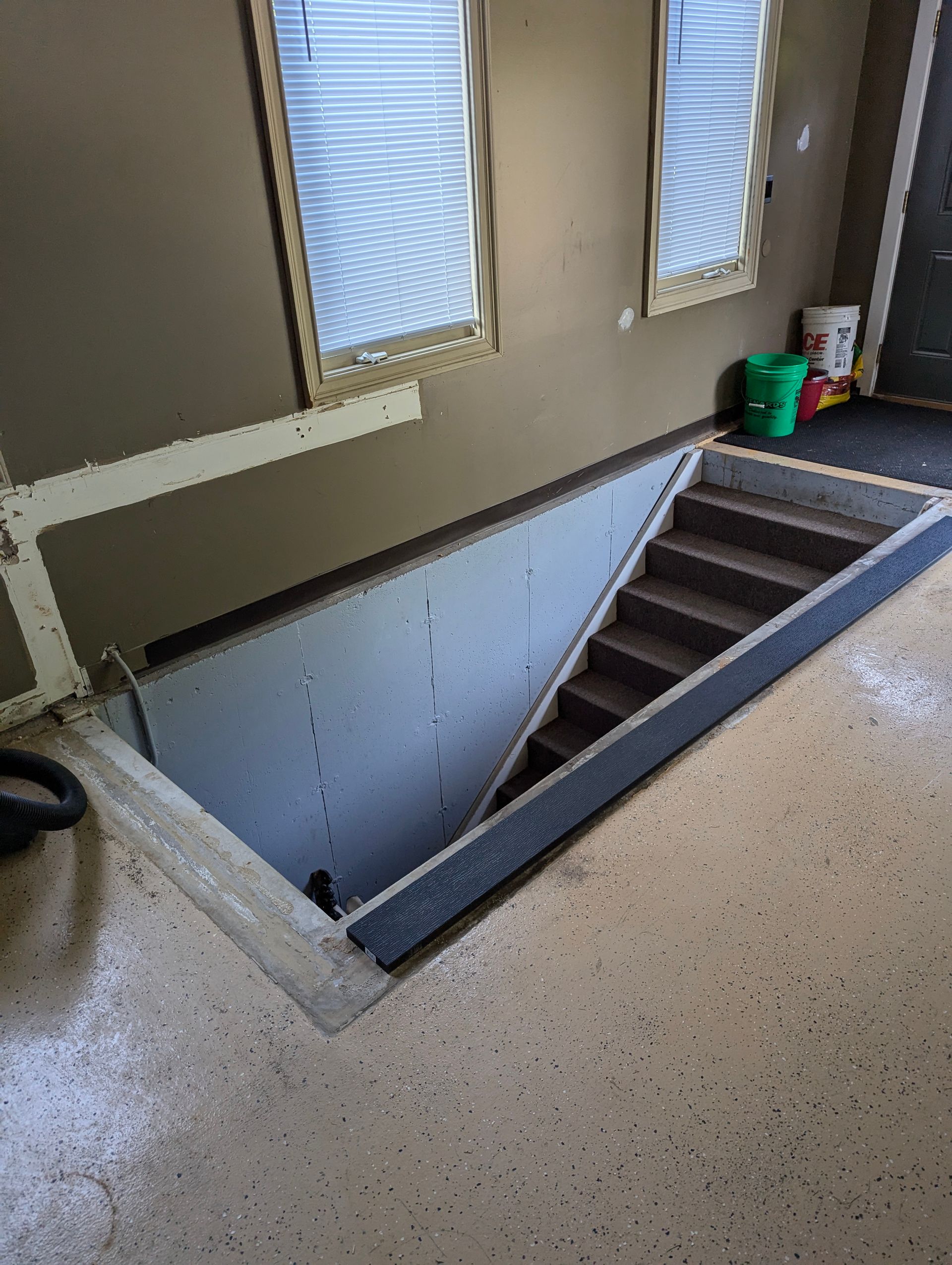 Staircase leading down to a basement, framed by a concrete opening in a speckled floor and walls with two windows.
