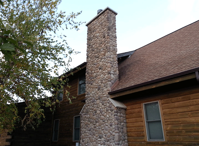 A tall stone chimney rises against the side of a brown log cabin home with a shingled roof under a clear sky.