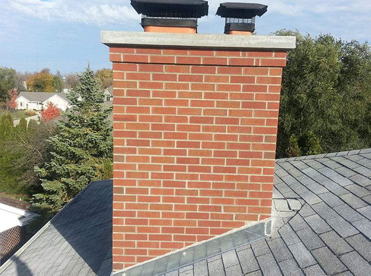 A red brick chimney with two chimney caps on a grey shingled roof with trees in the background.