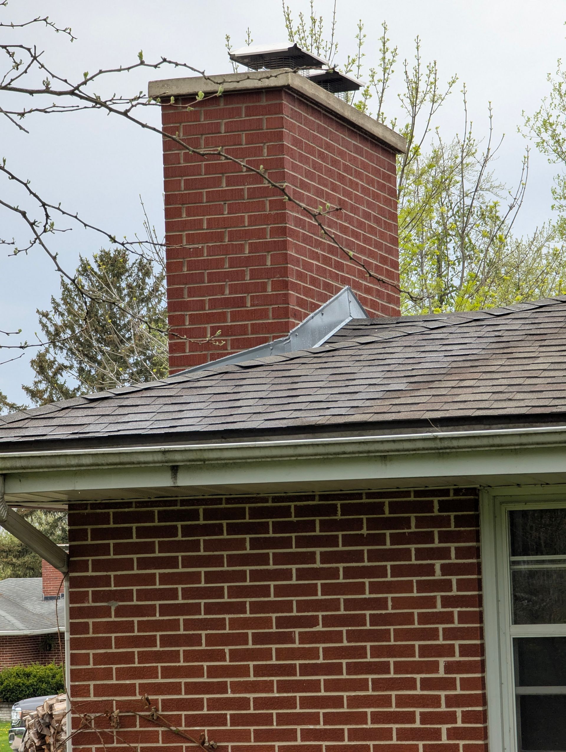 A red brick chimney rises from a shingled roof with metal flashing on a bright day.