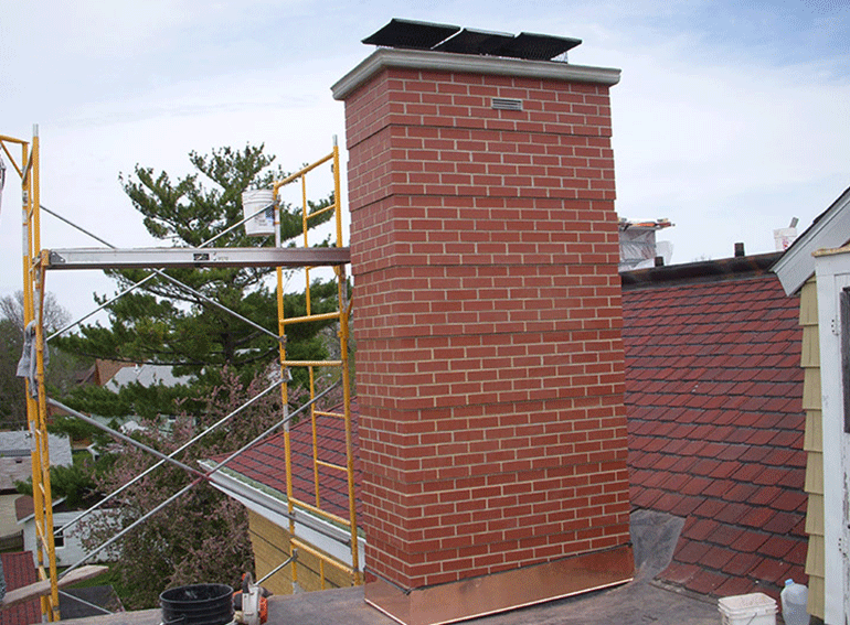 A red brick chimney with a metal cap sits on a roof next to yellow scaffolding under a cloudy sky.