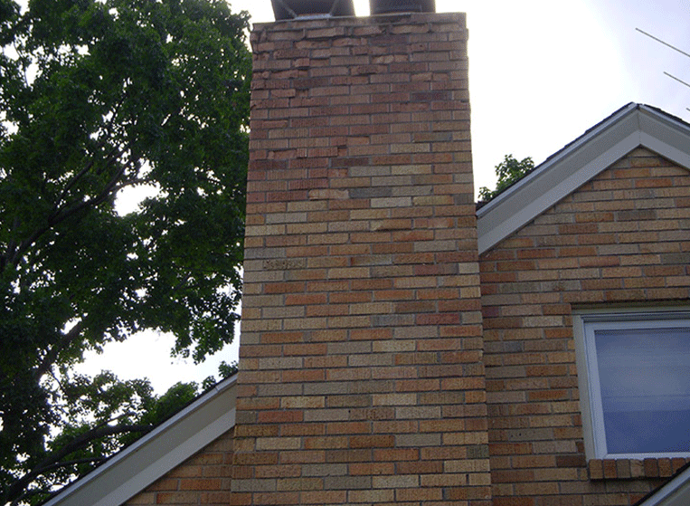 A tall brick chimney stands against the side of a house with light-colored brick siding near a large green tree.