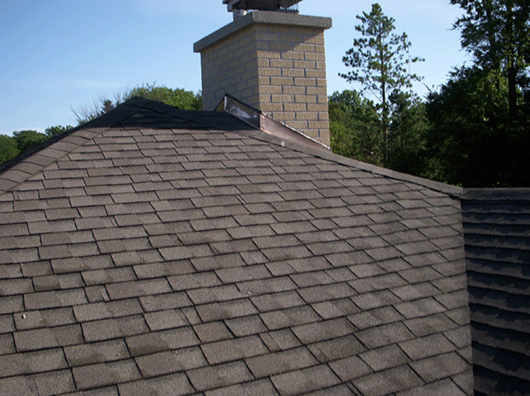 A view of a shingled residential roof with a tall brick chimney set against a blue sky and green trees.