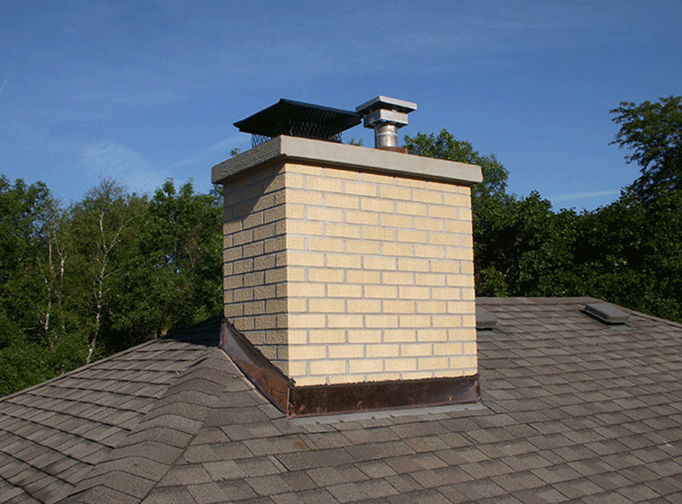 A light-colored brick chimney sits on a shingled roof under a clear blue sky, featuring a metal cap and copper flashing.