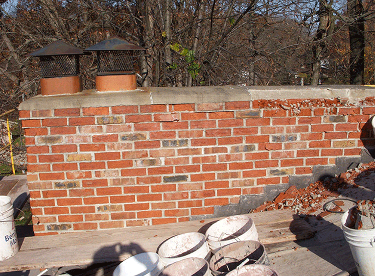 A brick chimney on a roof with two metal caps, viewed from scaffolding with several white buckets nearby.