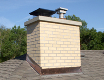 A brick chimney with a metal cap and a vent pipe, situated on a shingled roof against a blue sky.