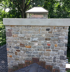 A stone chimney with a concrete cap and a metal chimney cover, situated on a dark shingled roof against a background of trees.