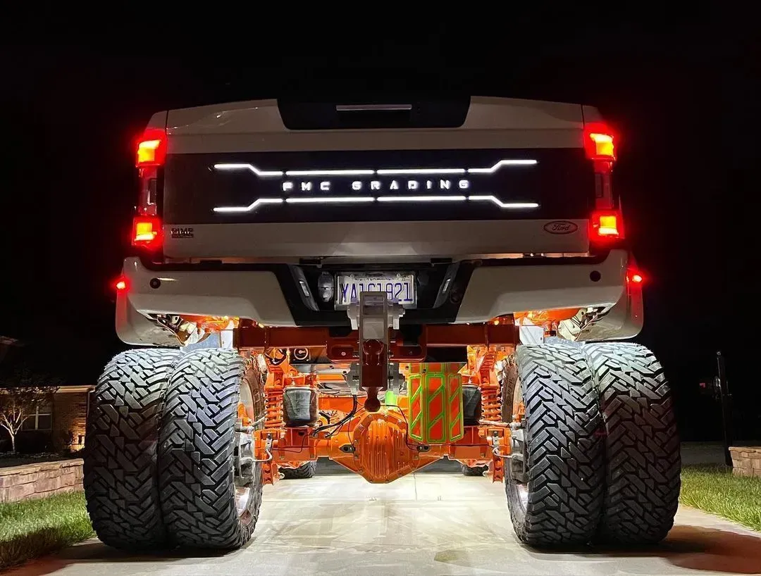 Rear view of a white, lifted truck with dual rear tires lit up with orange underglow and a custom illuminated tailgate.
