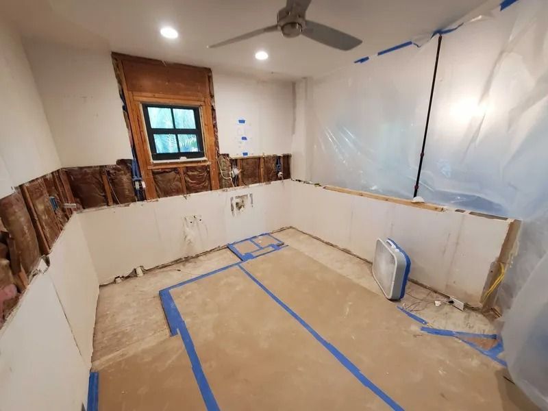 Room under renovation: exposed wall studs, drywall, plastic sheeting, window, ceiling fan. Blue tape on floor.