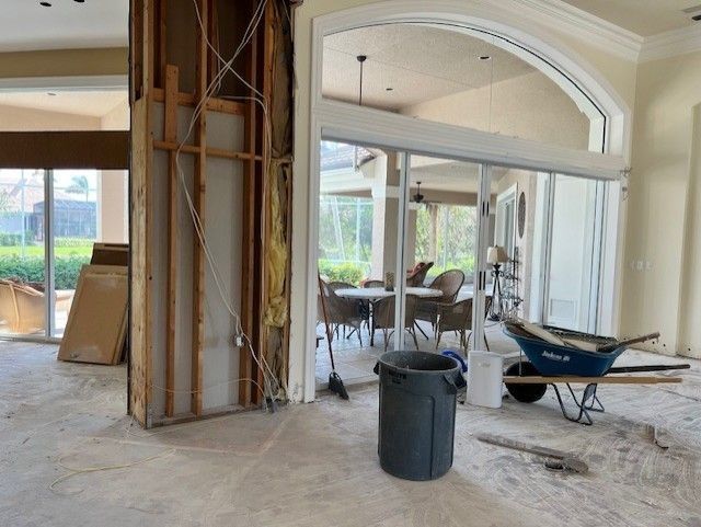 Interior renovation with exposed wall studs, wires, and debris; adjacent to a room with glass doors and outdoor dining.