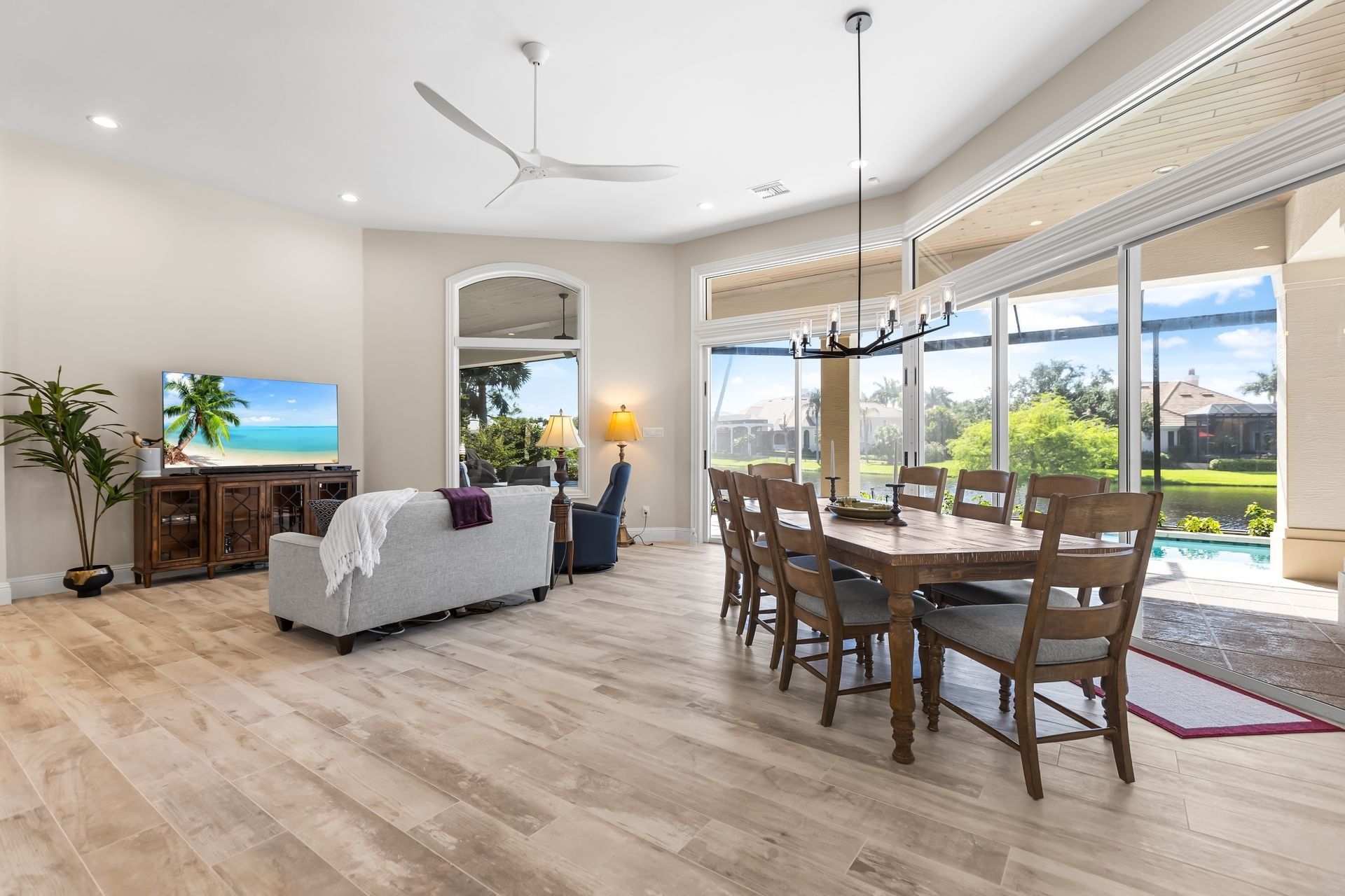 Spacious living area with dining table, TV, and large windows overlooking a pool and lush greenery.