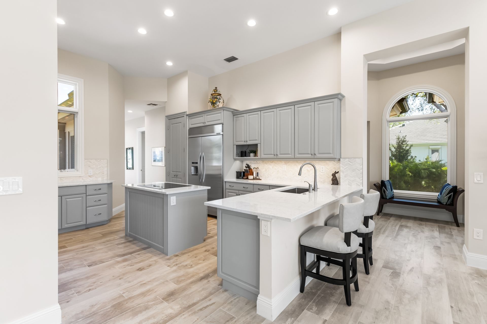 Light gray kitchen with island, cabinets, white countertops, bar stools, and arched window.