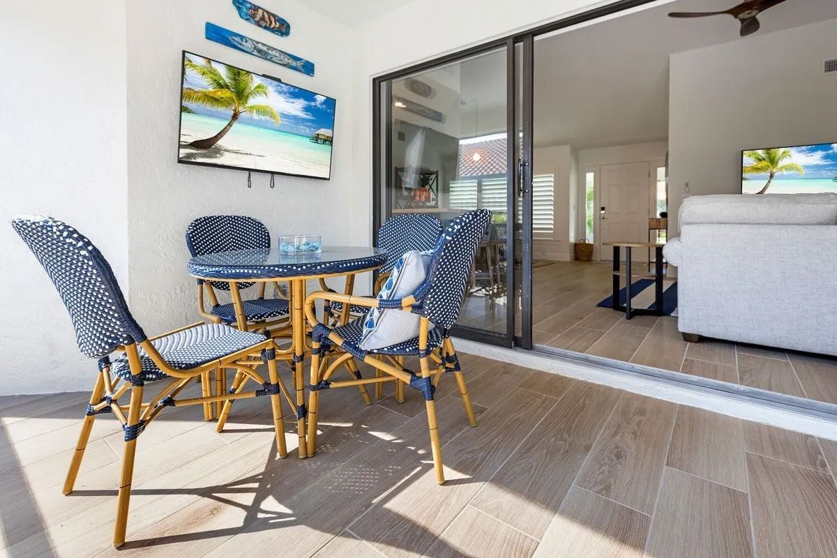 Outdoor dining set on a patio with a TV showing a beach scene, near a sliding door to the interior.