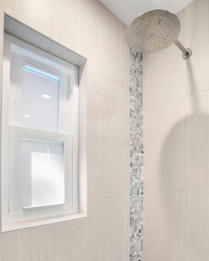 Shower with a window. White and gray tiled walls with a mosaic strip. Large shower head.