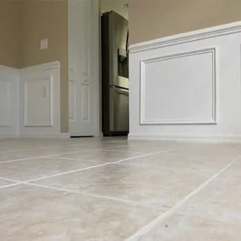 Low-angle view of beige tiled floor, white wainscoting, and stainless steel refrigerator in a room.