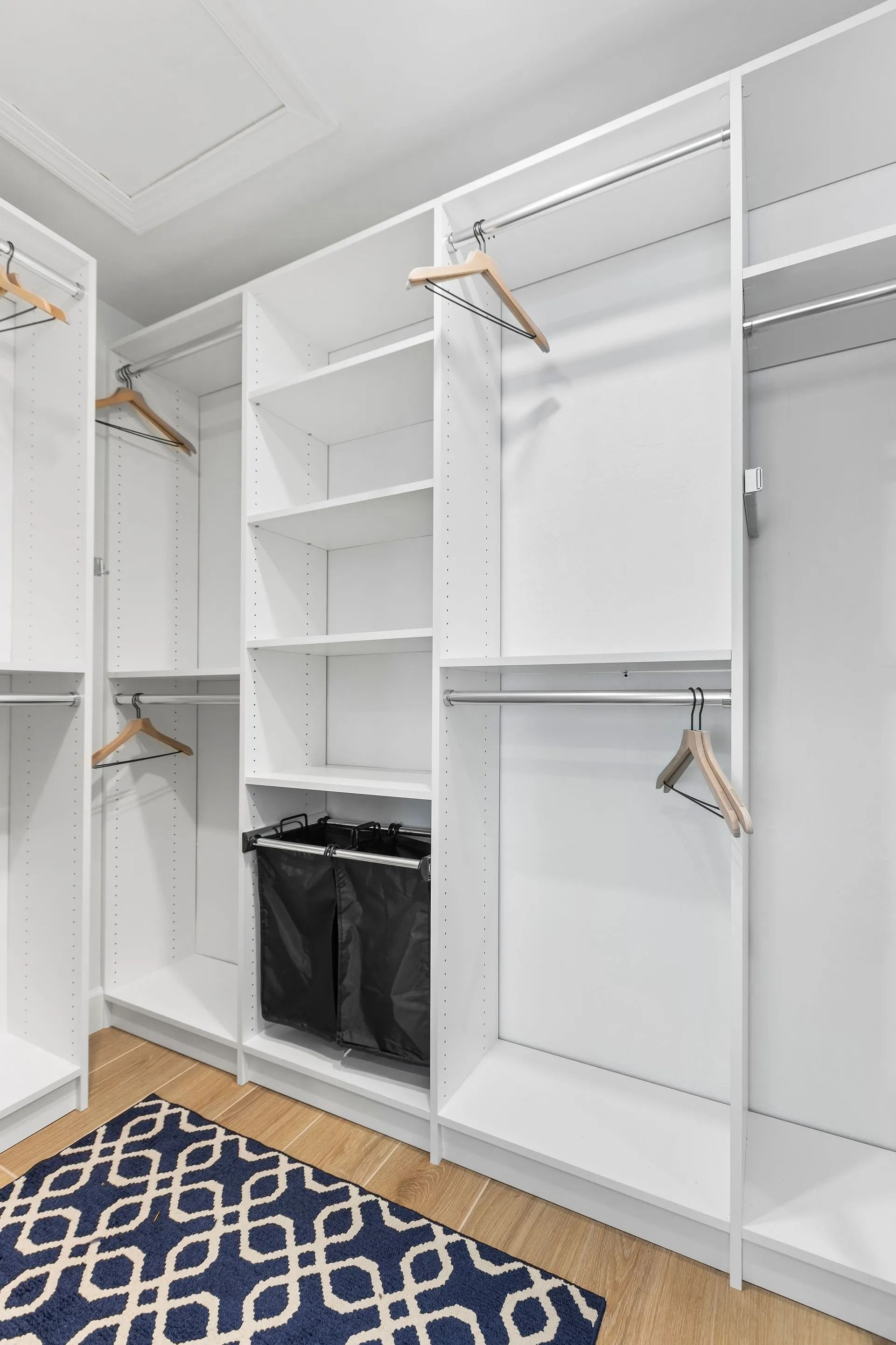 White closet with shelves, hanging rods, and a black hamper. A blue and white patterned rug is in front.