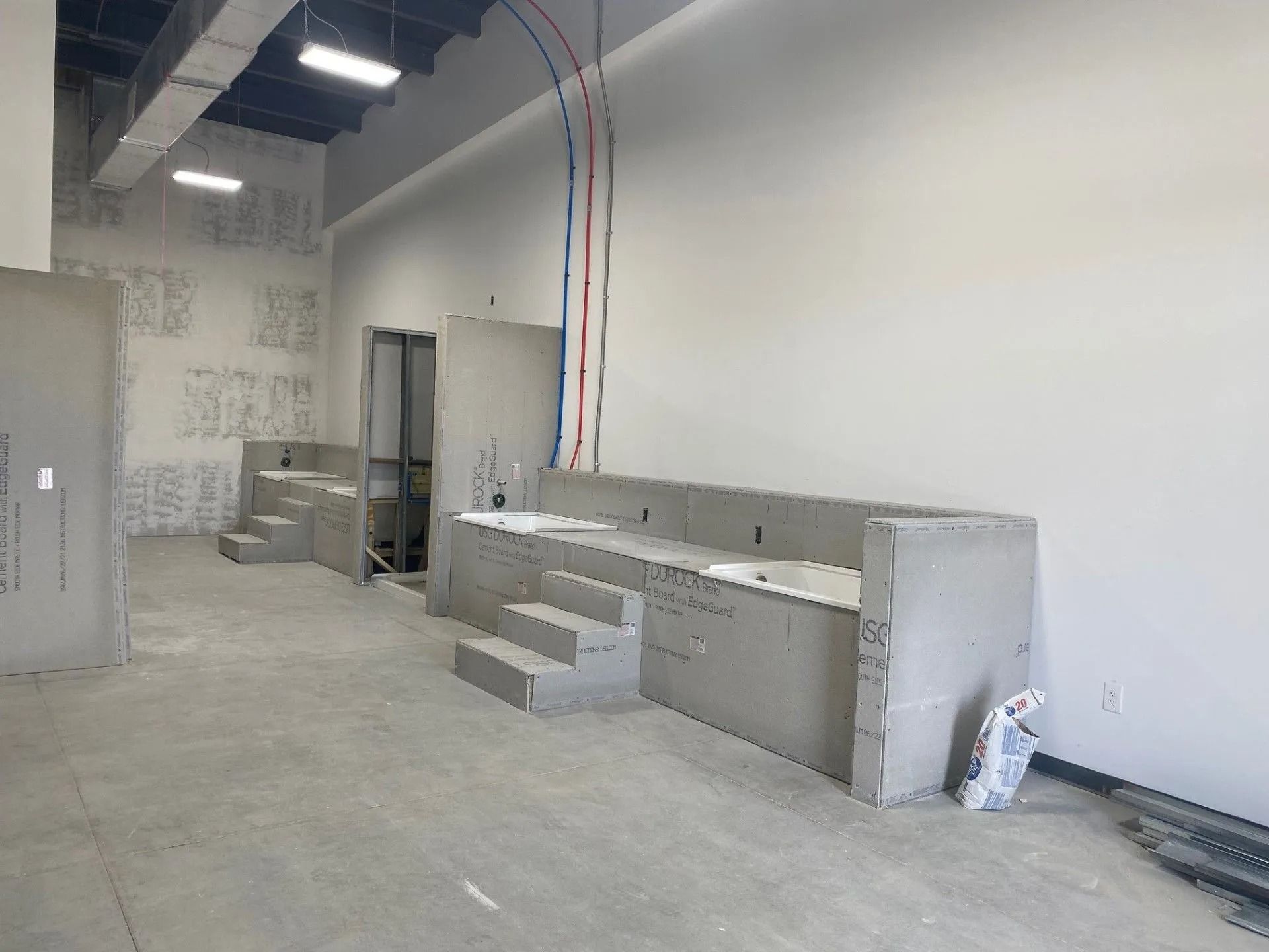 Interior construction site with gray concrete and drywall. Plumbing visible on the wall. Steps and countertops are partially built.