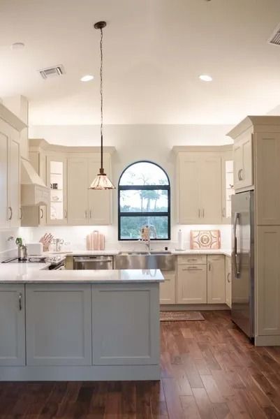 Cream-colored kitchen with island and arched window. Stainless steel appliances, hardwood floors, and overhead lighting.