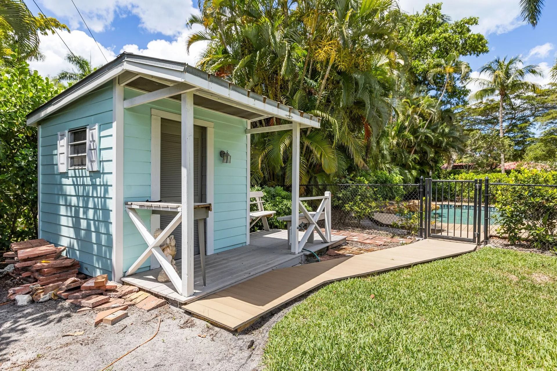 Small blue shed with porch in a yard, near a pool, surrounded by greenery.