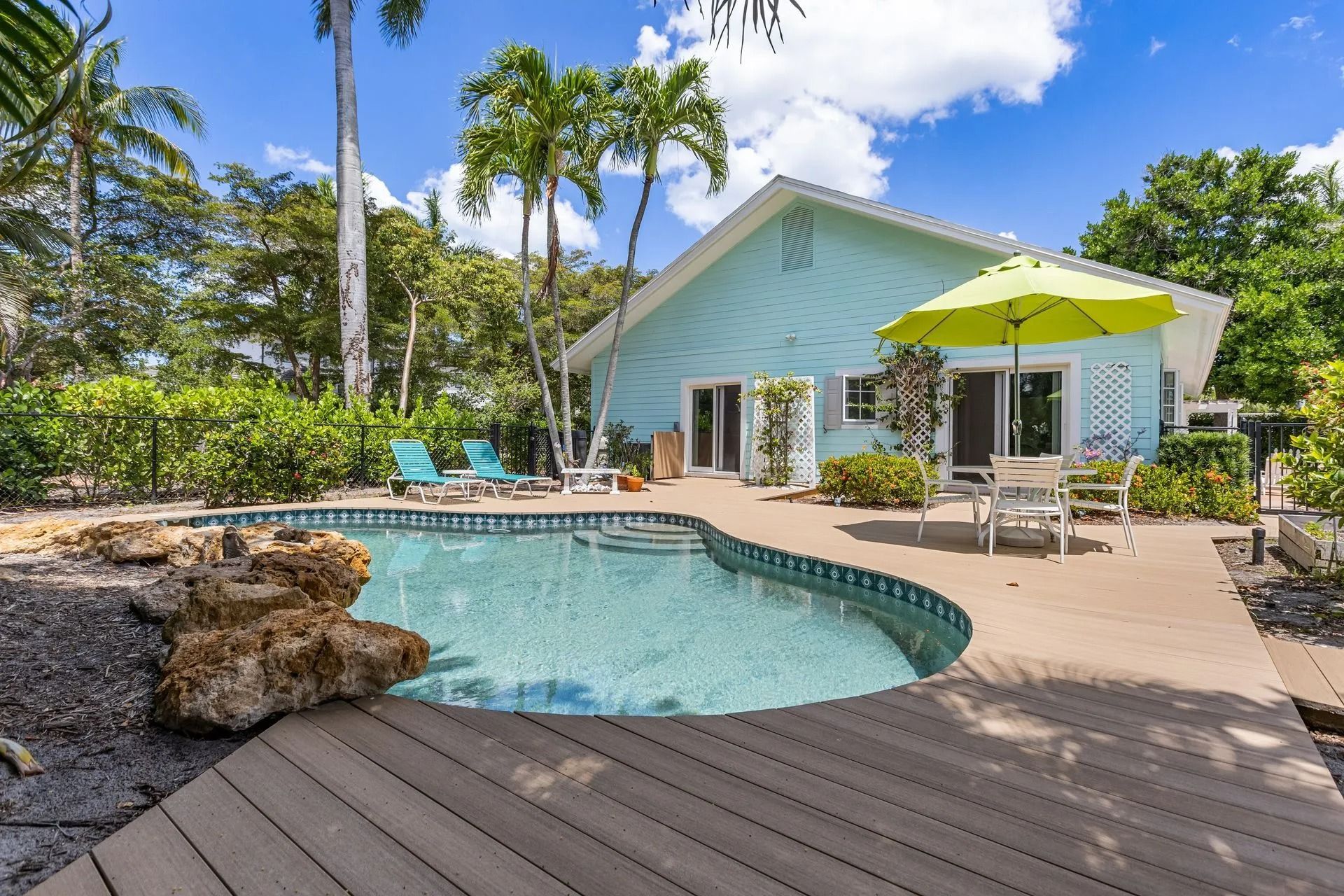 A turquoise house with a pool and deck surrounded by lush greenery under a sunny sky.