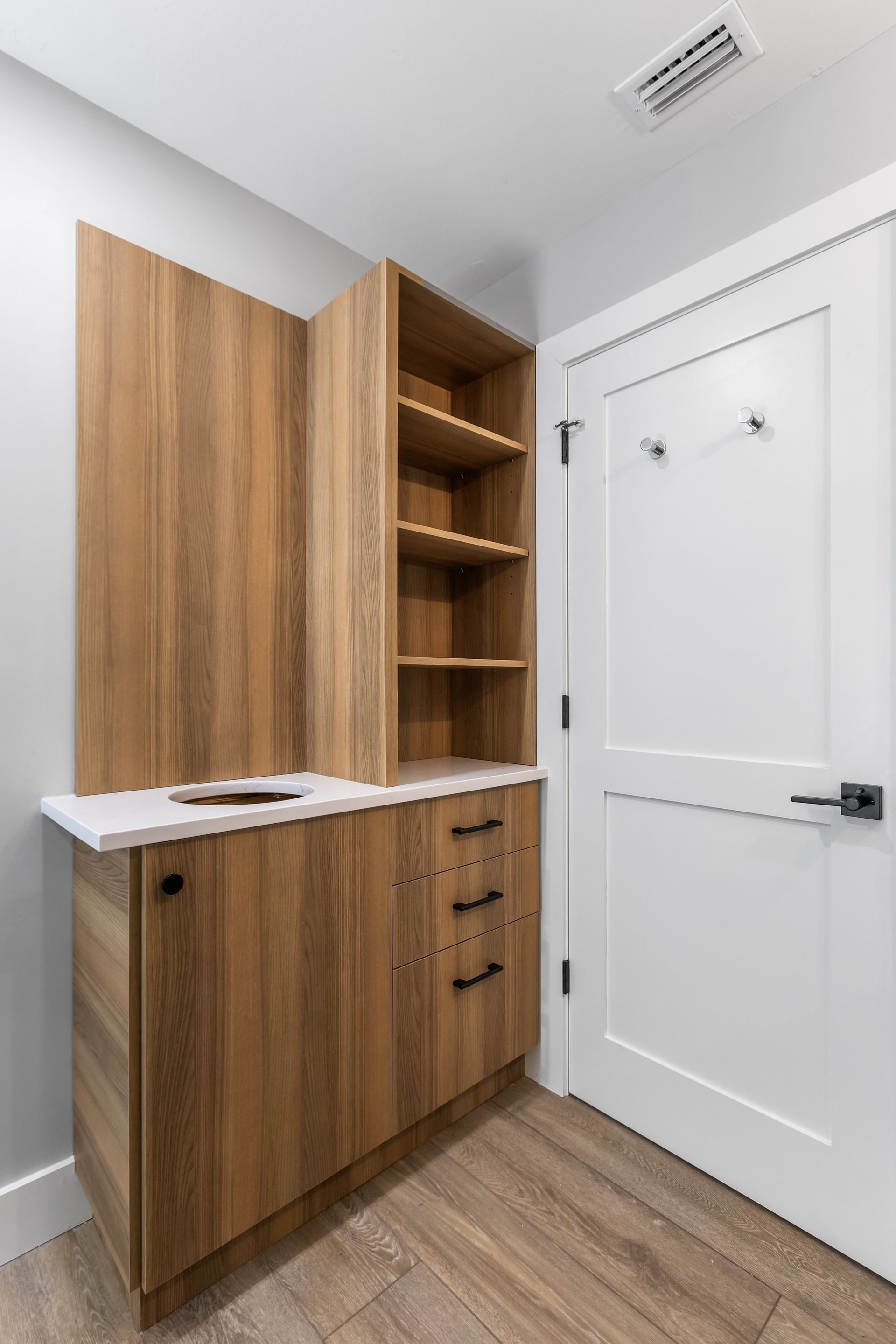 Wooden cabinet with open shelves, drawers, and white countertop next to a white door with coat hooks.