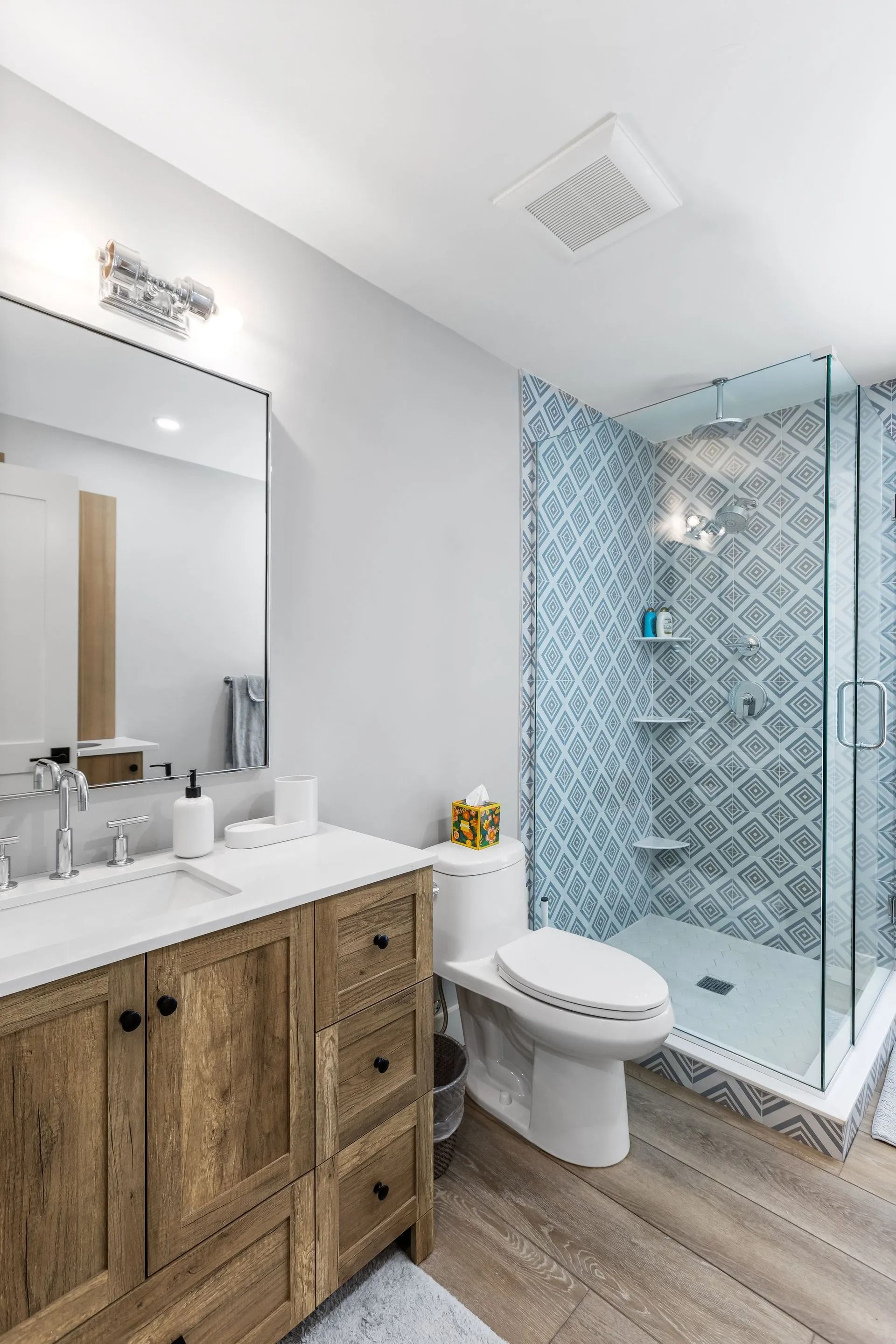 Bathroom with wooden vanity, blue patterned shower, and toilet on wood-look floor.