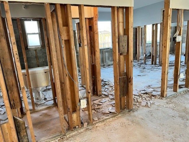 Interior framing of a room stripped of walls, revealing studs and debris. A window and fixtures are visible.