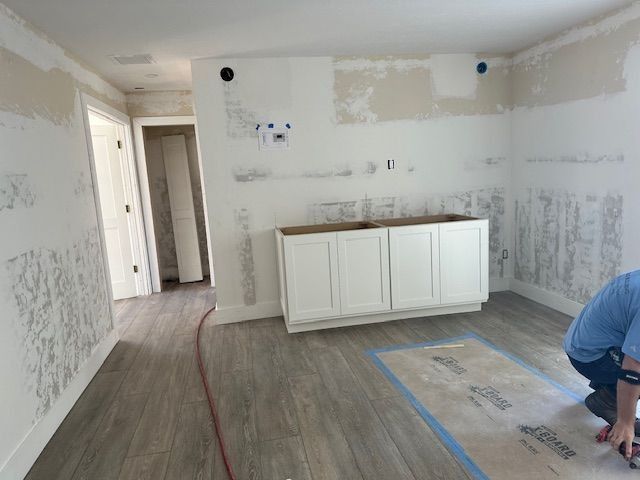 Room under renovation; white cabinets installed. Gray flooring, damaged walls, a person working.