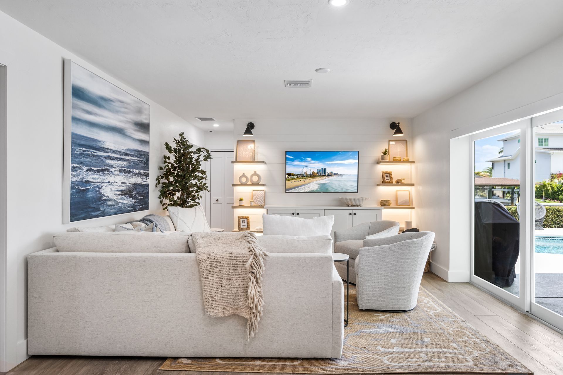 Bright living room with a white couch, a rug, and a TV on a white wall, with doors to a pool.