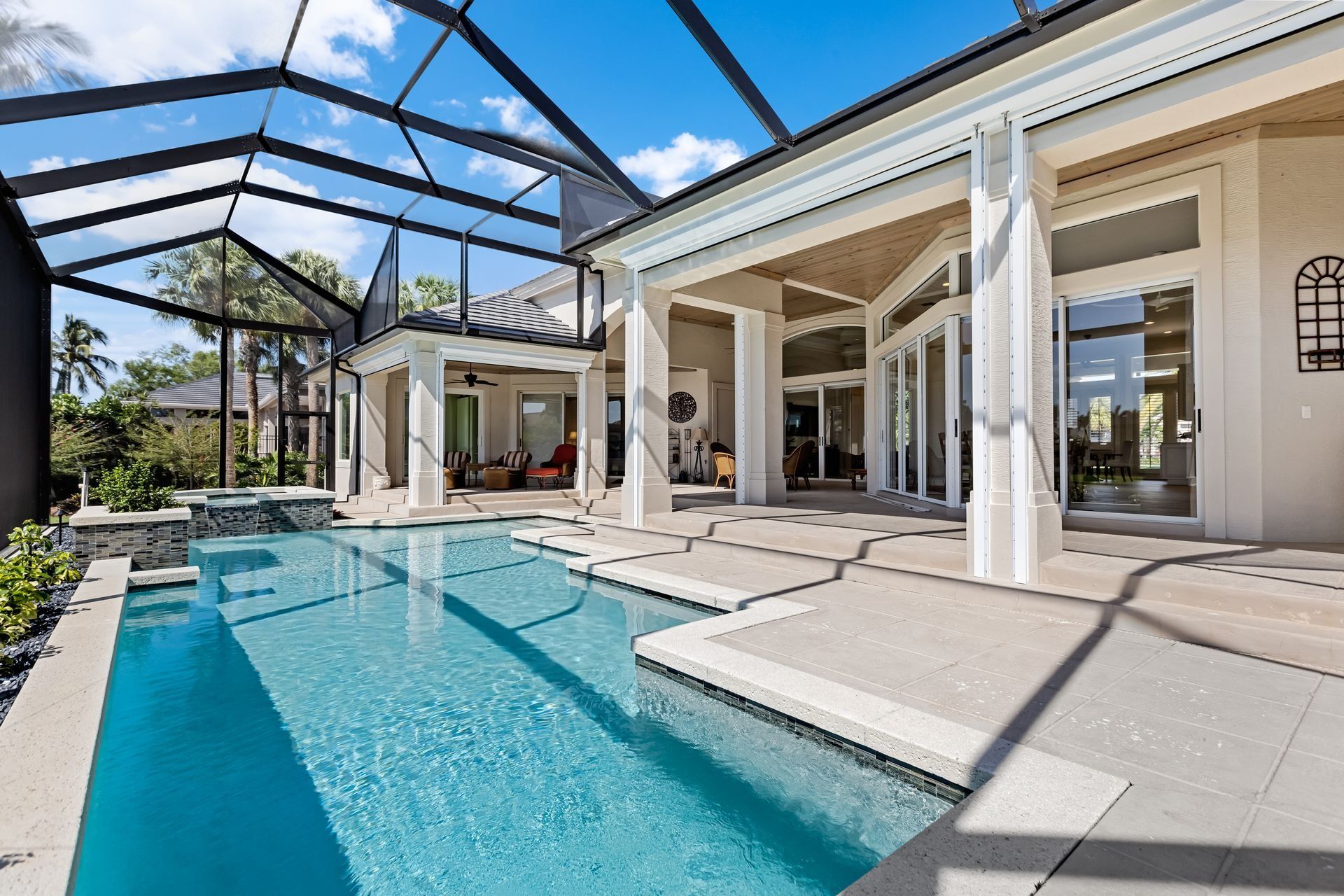 Pool and patio of a home. Blue pool with steps and a screened enclosure. White columns and building. Sunny day.