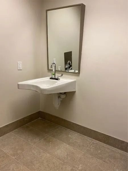 White wall-mounted sink with faucet, mirror, and soap dispenser in a corner; beige floor and walls.