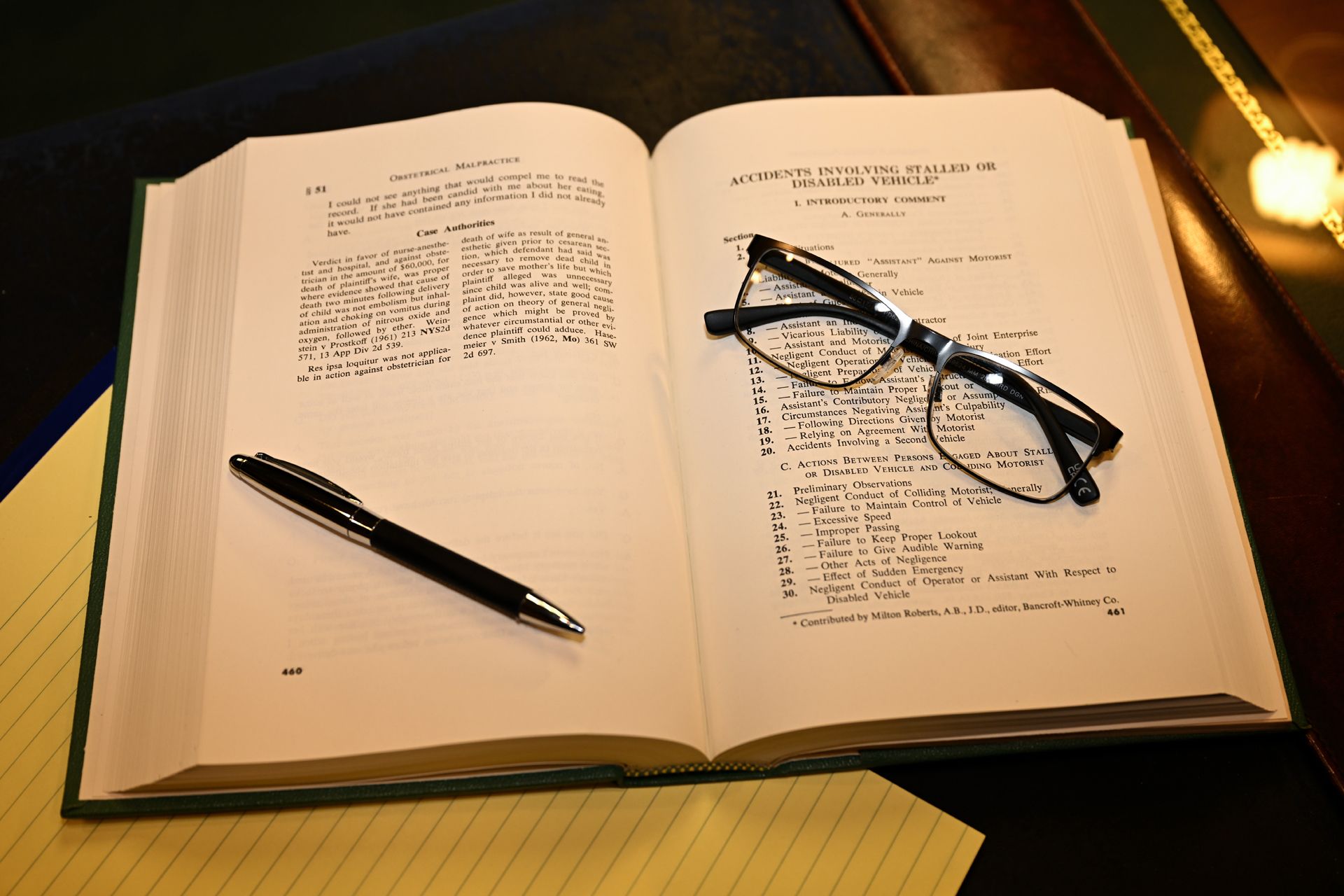 An open book on a dark surface with a black pen and a pair of reading glasses resting on the pages.
