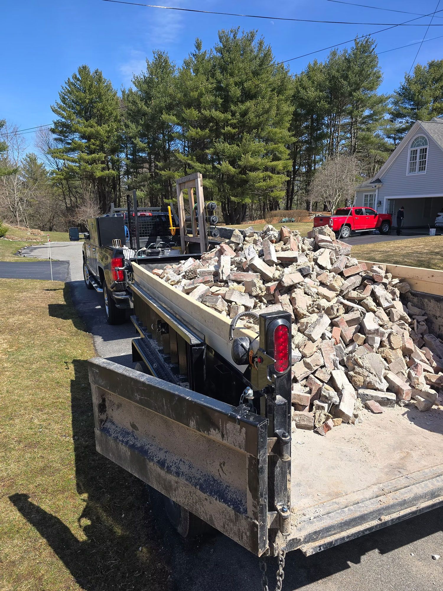 Dump trailer full of broken concrete beside a road, with trees and a red pickup in the background.