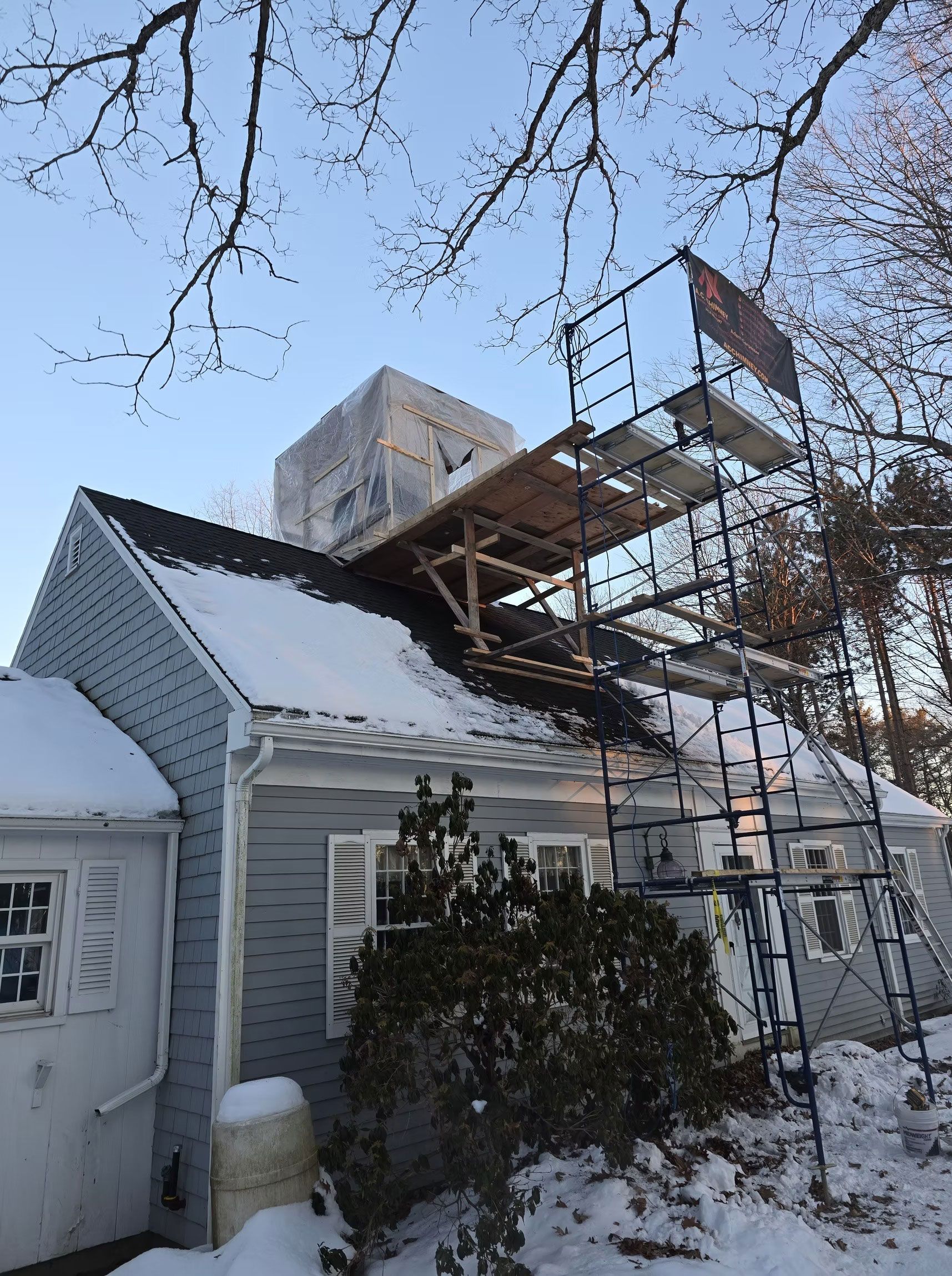 Snow-covered house with scaffolding on the roof and bare trees overhead