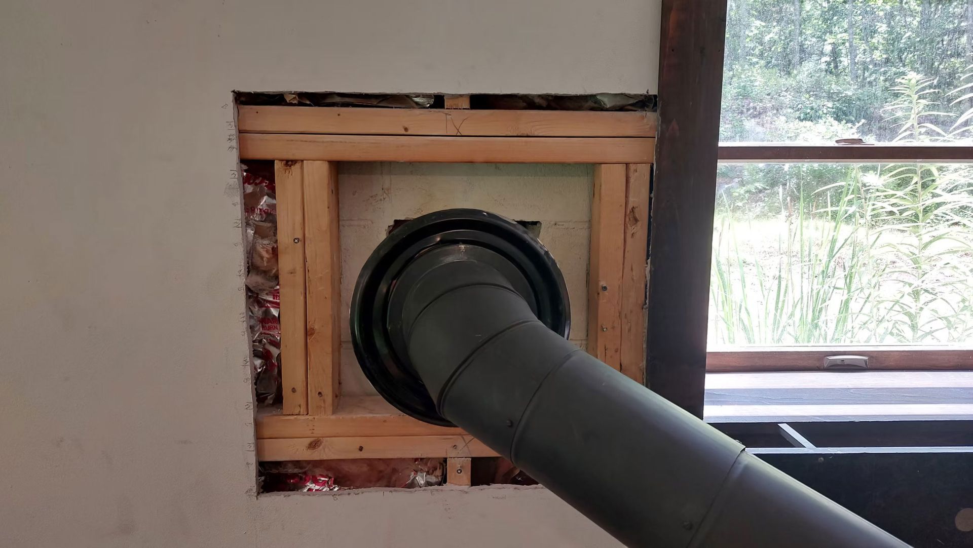 Black pipe extending through a wooden-framed wall opening beside a window.