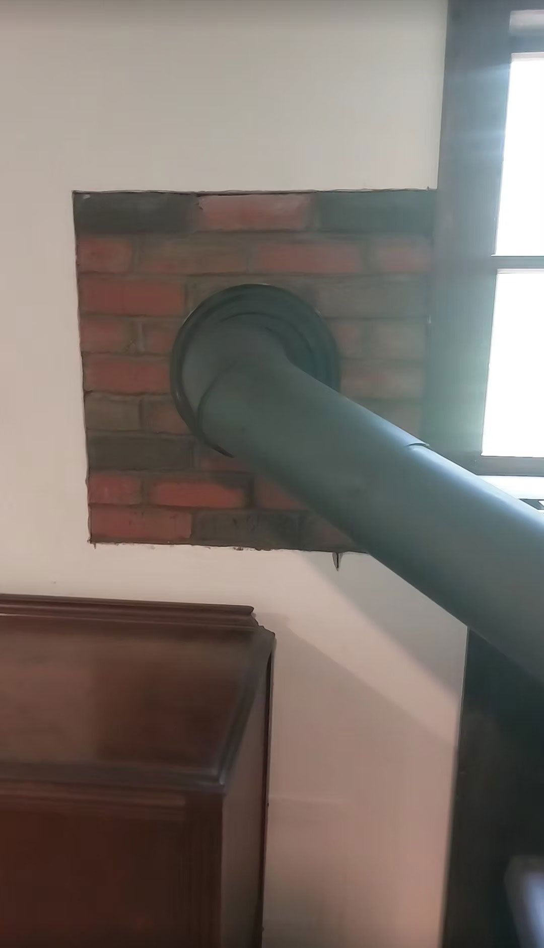Gray stove pipe extending through a brick wall near a window and wooden furniture