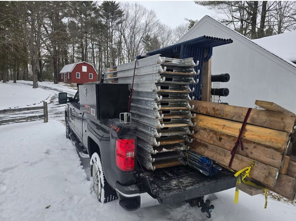 Black pickup truck hauling stacked metal panels and wooden boards in a snowy yard near a red barn
