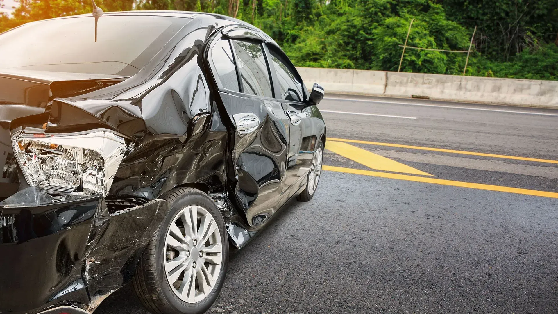 Severely damaged black car on a wet road after a crash, with dents along the side and front.