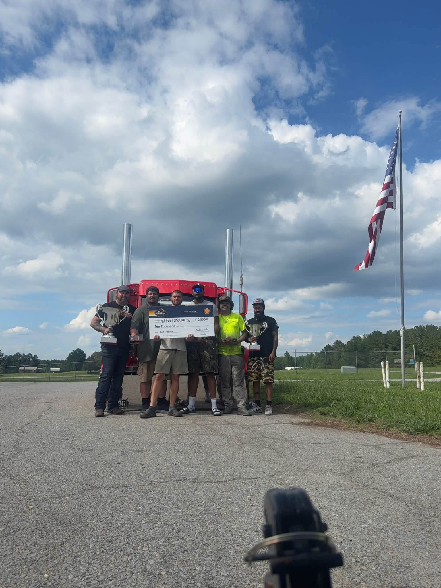 Group of people holding a sign on a gravel road under cloudy sky near a large American flag.