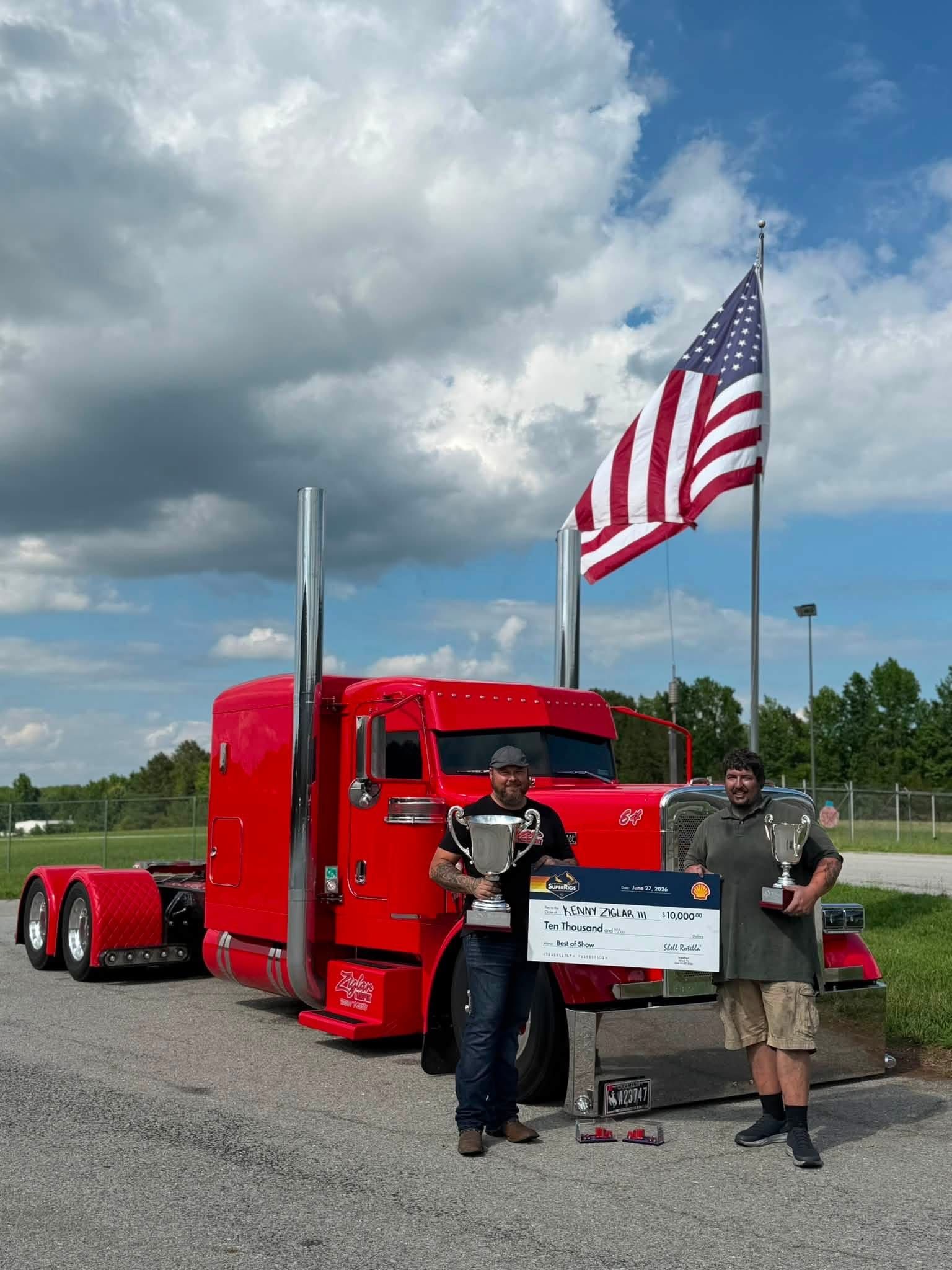 People posing by a red semi truck with an American flag in a gravel lot.