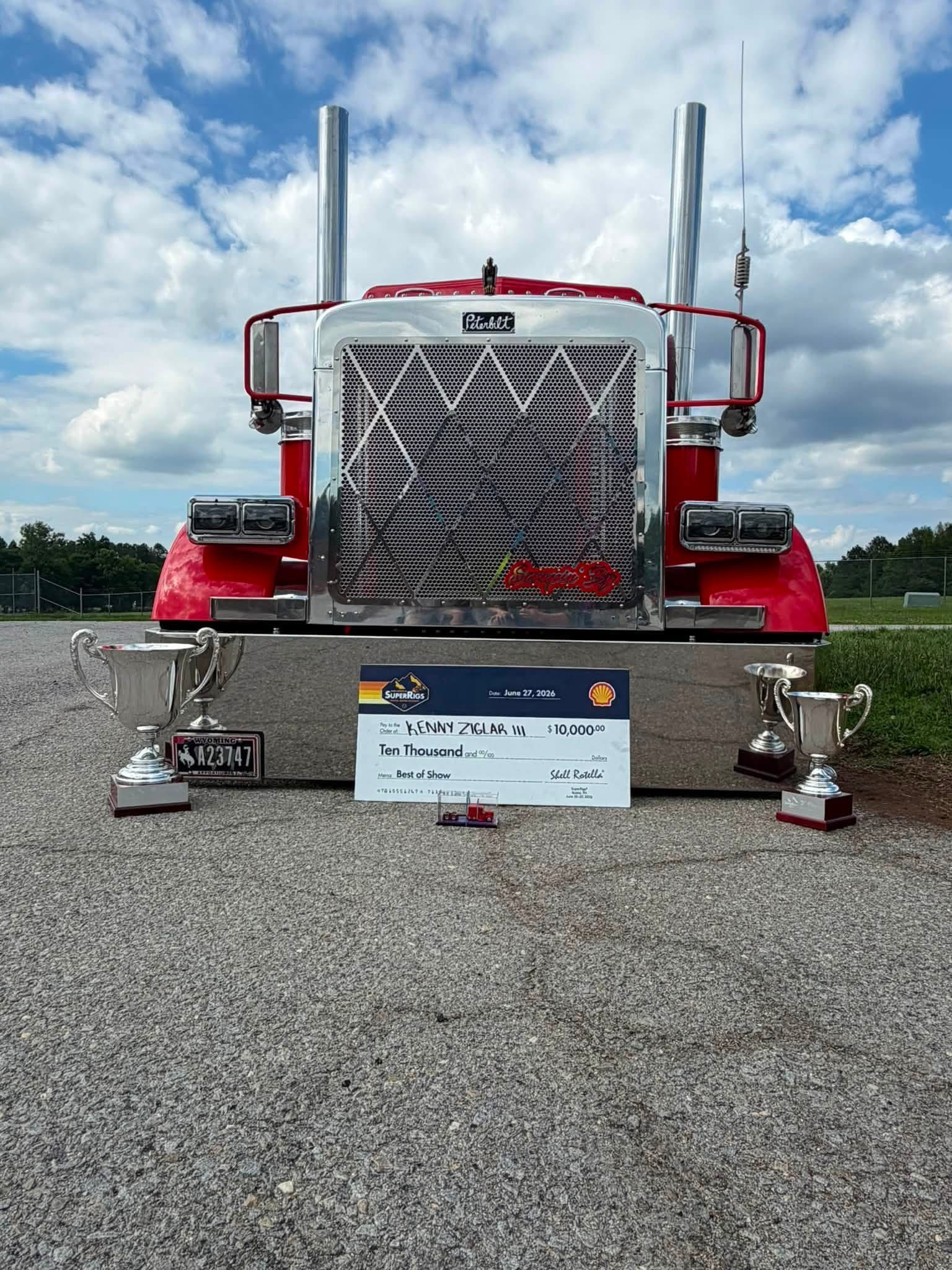 Red semi-truck on a gravel lot with a trophy and award plaque in front under a cloudy sky