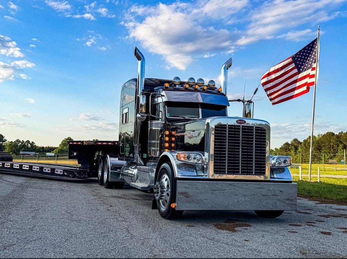 Black semi truck with chrome details parked on gravel beside a large American flag at sunset