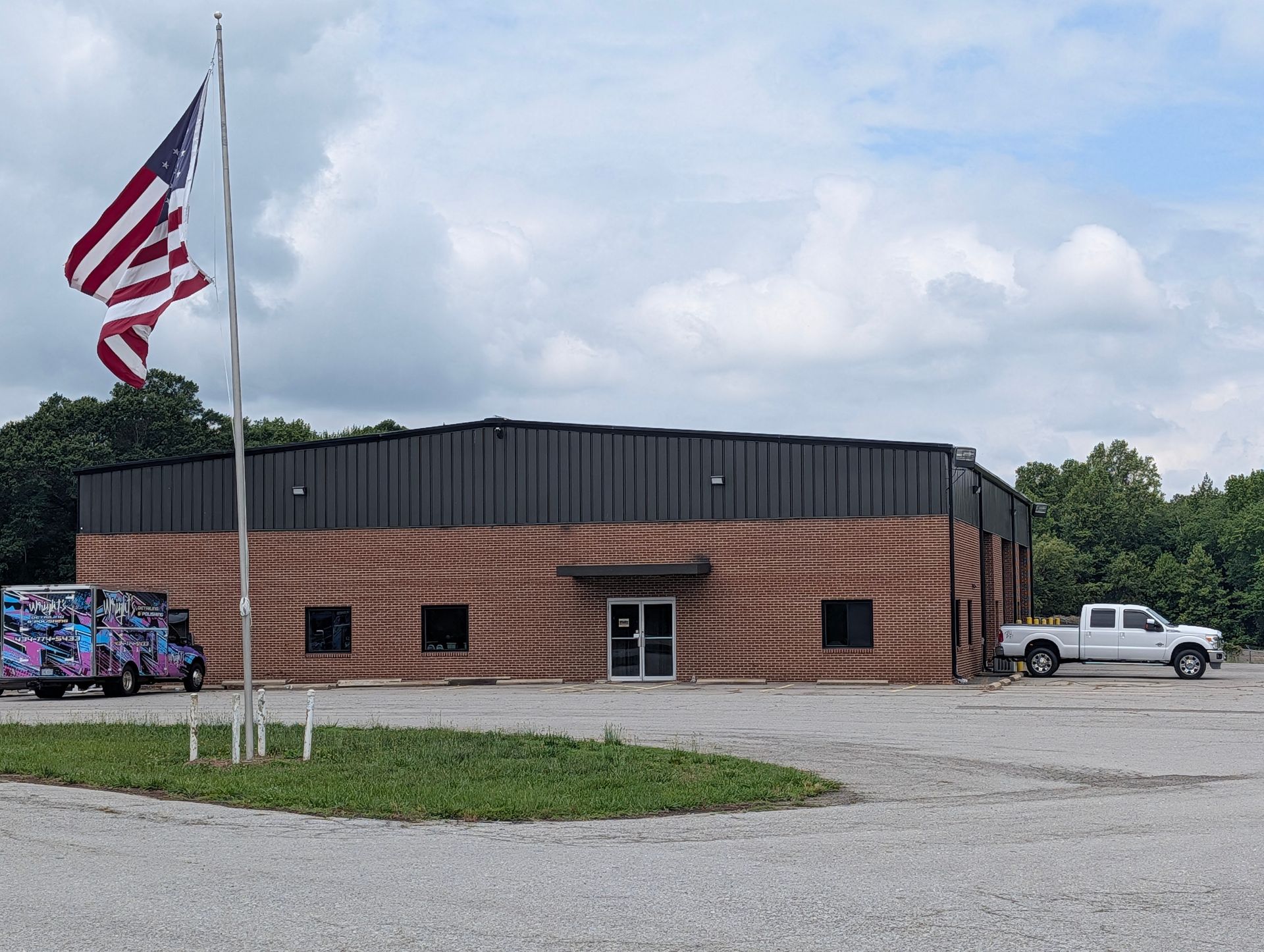 Brick building with a black roof, U.S. flag, and parked truck beside a gravel lot under cloudy sky