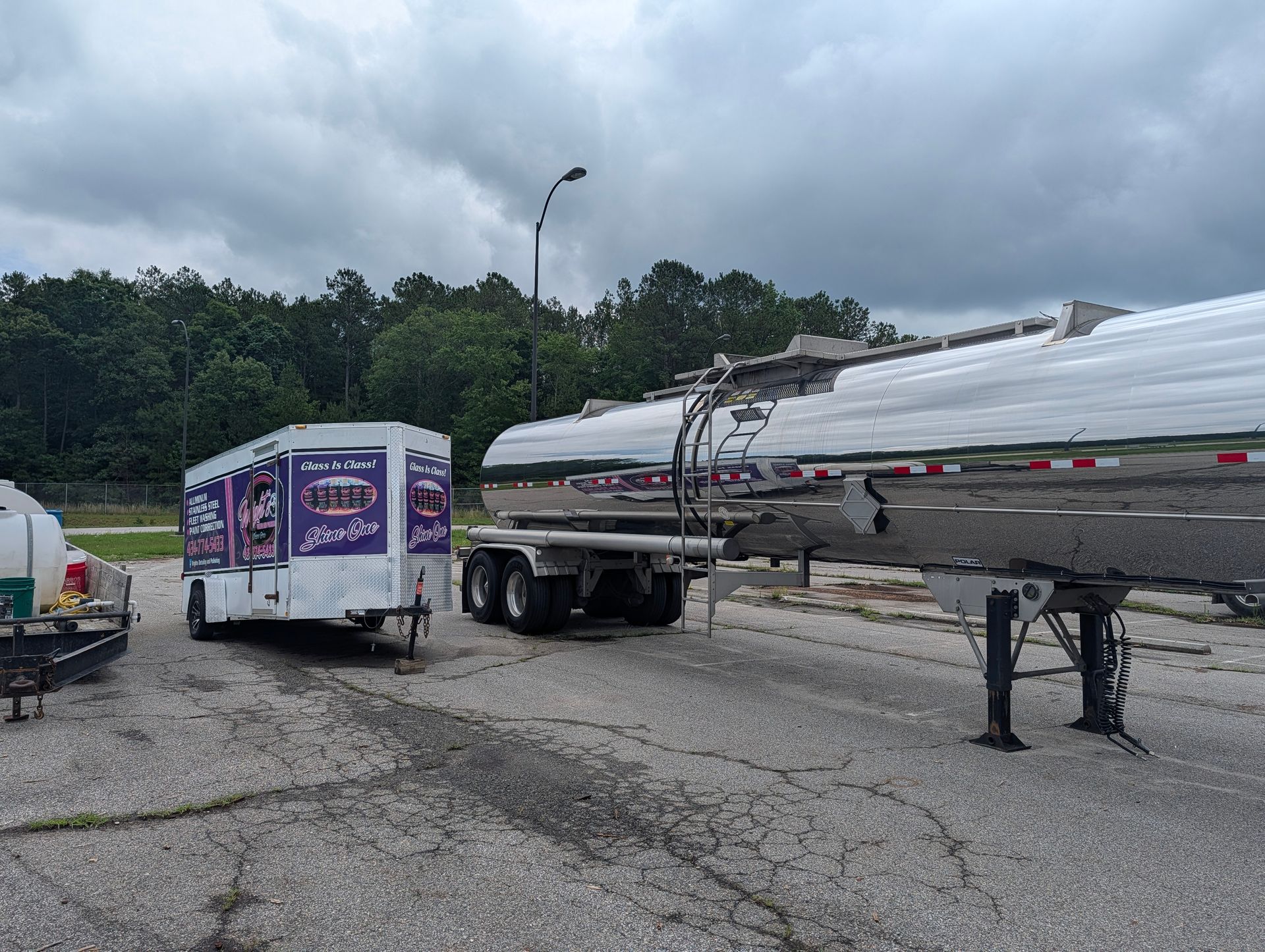 A tanker truck and purple box truck parked on a cracked lot under cloudy skies, with workers nearby.