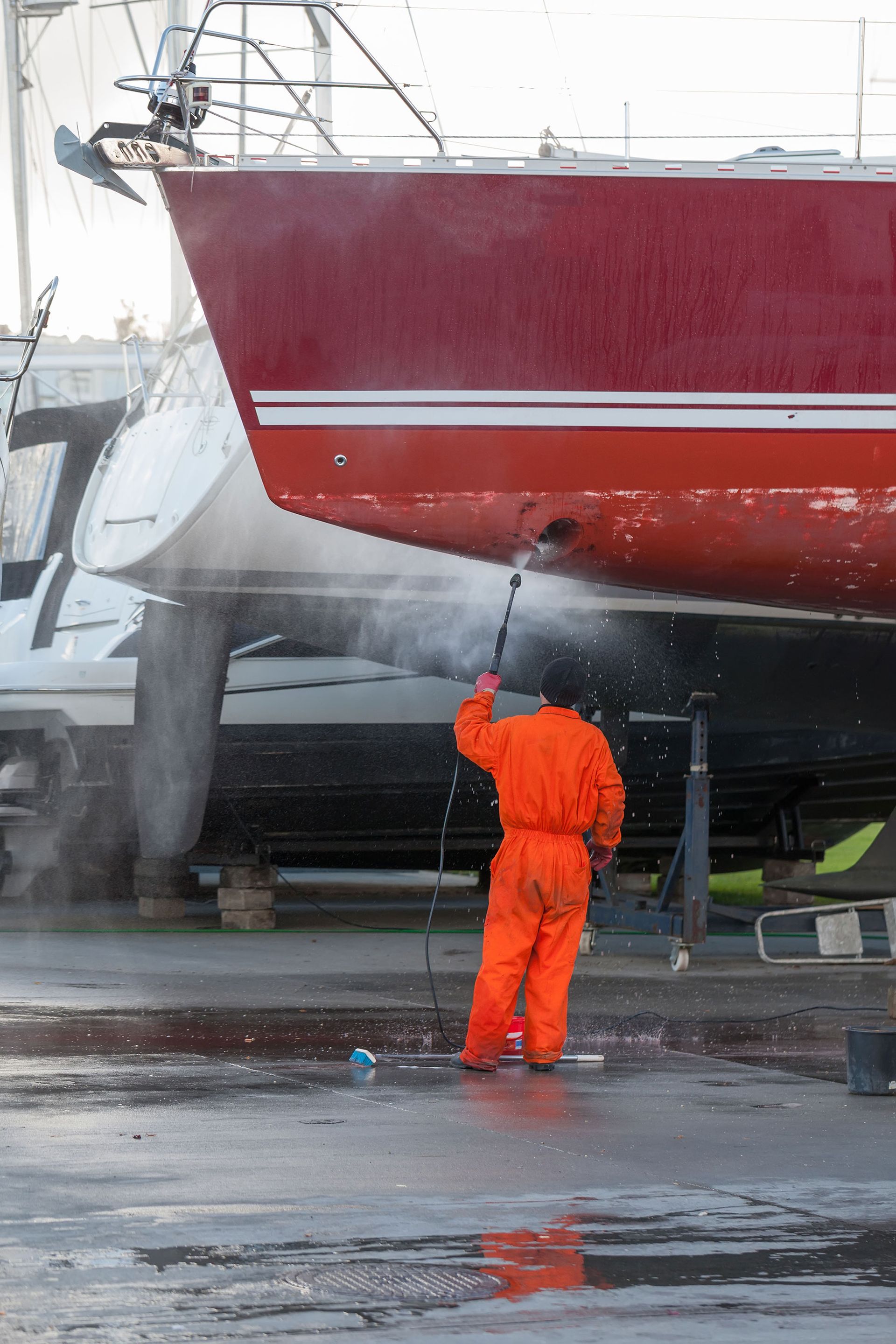 Person in orange coveralls pressure-washes the hull of a red boat on land.