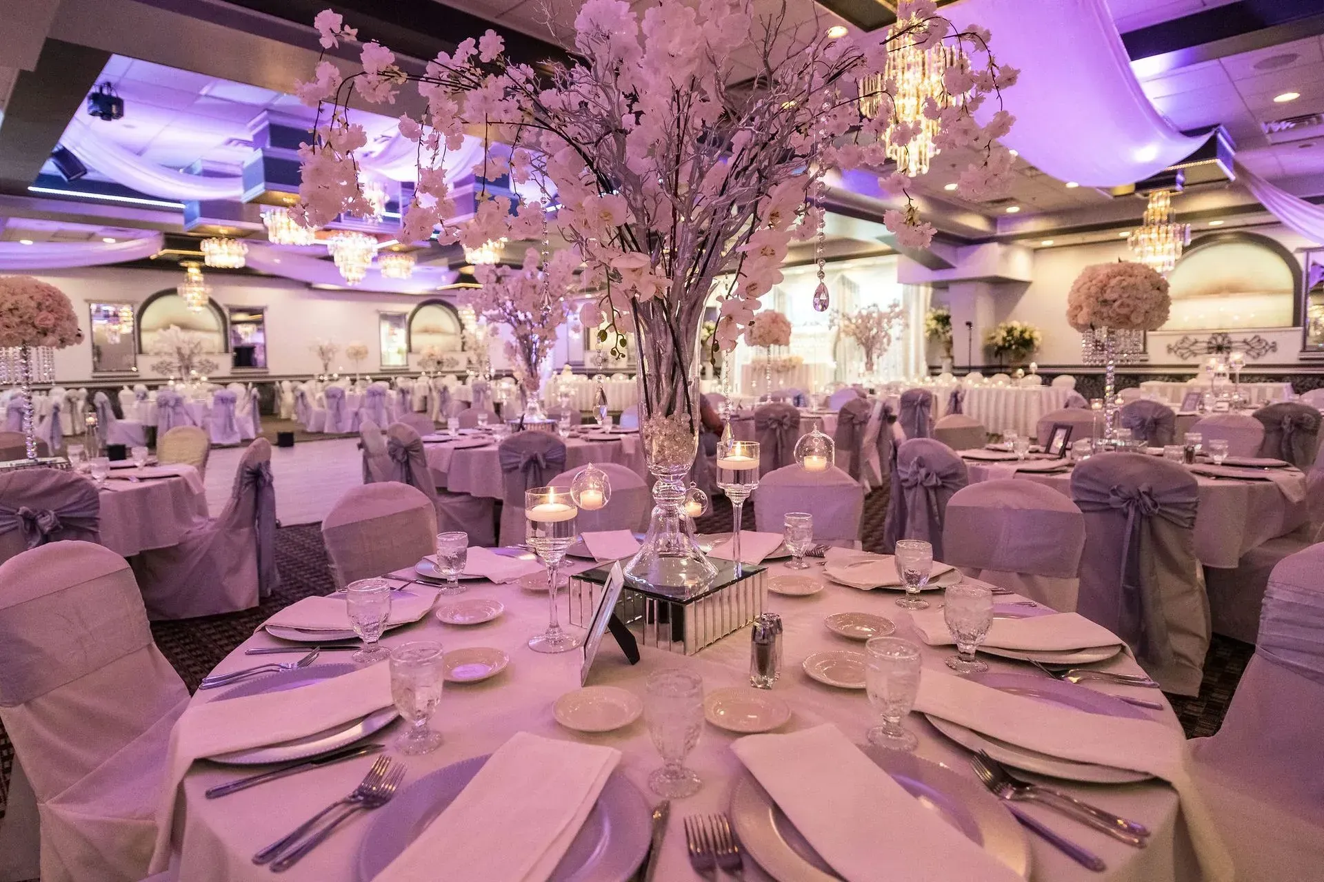 Elegant banquet hall with pink cherry blossom trees, round tables set with white linens, and soft purple lighting
