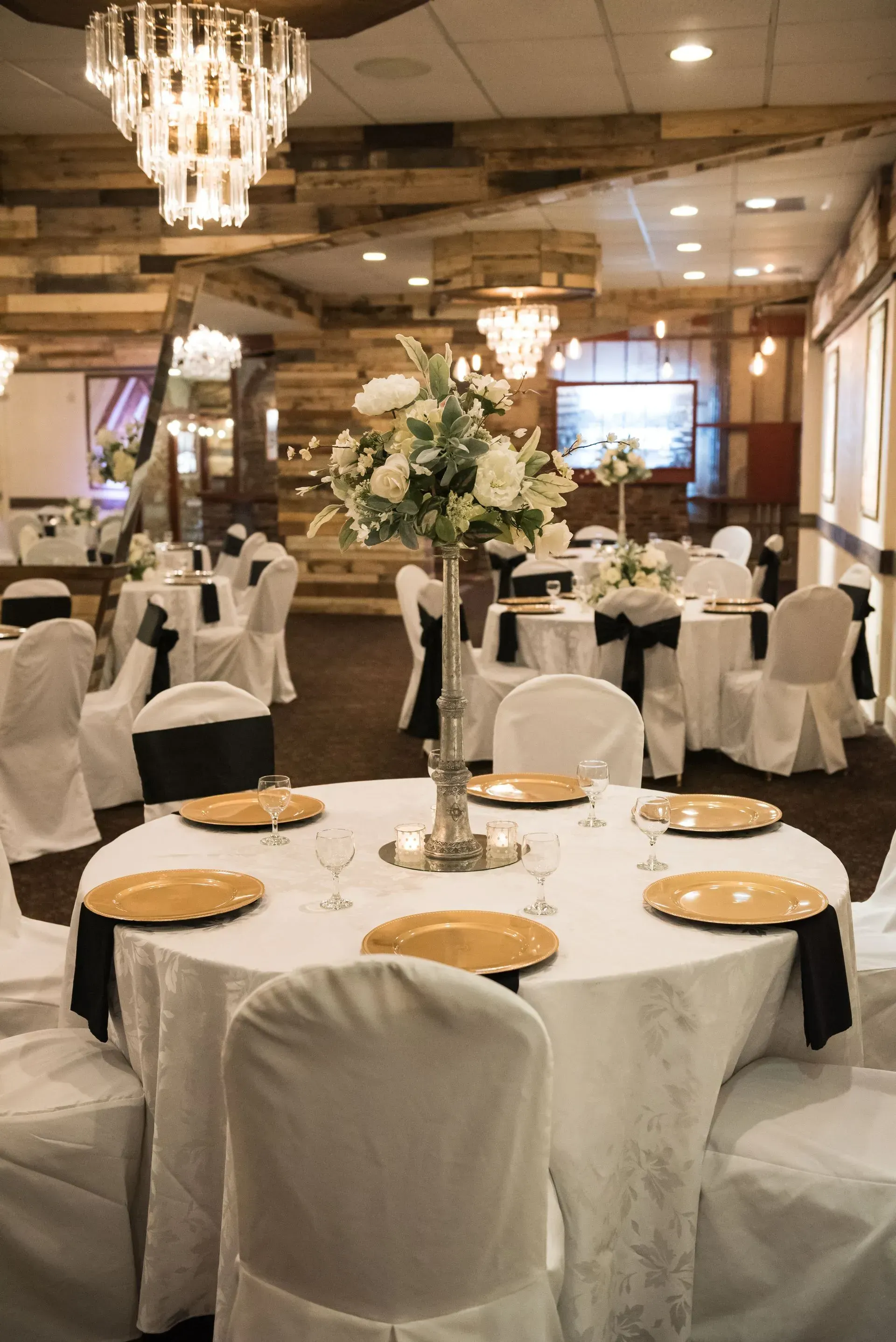 Banquet hall with round tables, white chair covers, chandeliers, and gold chargers set for an event